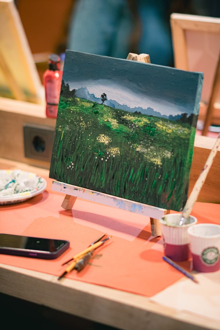 Close-up view of artistic landscape painting on easel surrounded by paintbrushes and a smartphone.